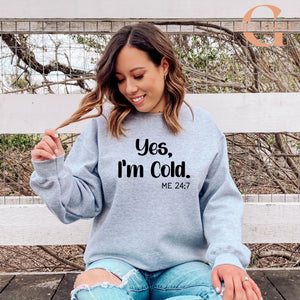 A woman wearing a gray sweatshirt with the text "Yes, I'm Cold. ME 24.7" is sitting on a bench in front of a fence.