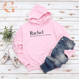 A pink hoodie with the word "Rachel" printed on it, along with a pair of jeans and sneakers, all laid out on a wooden surface.