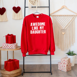 A red sweatshirt with the text "AWESOME LIKE MY DAUGHTER" is displayed on a black hanger, surrounded by red gift boxes and a beaded wall decoration.
