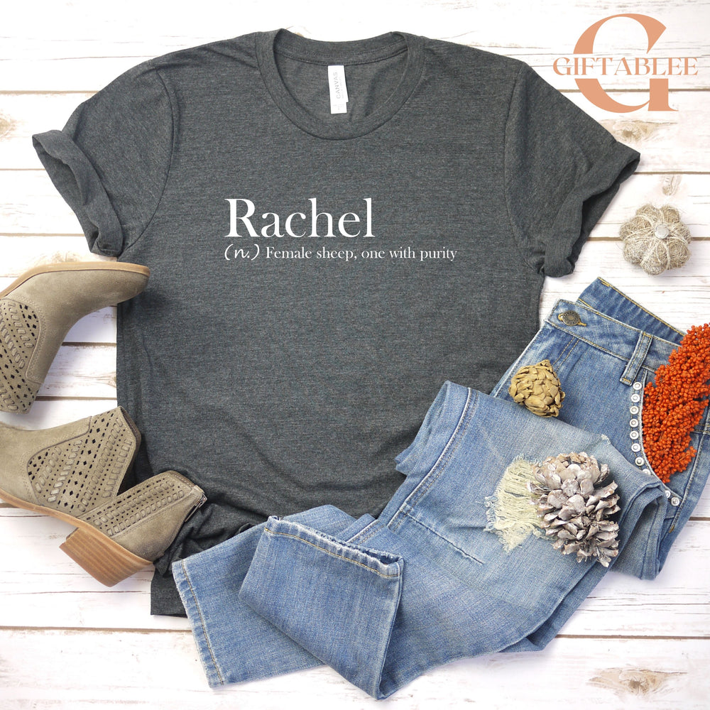 a casual outfit consisting of a gray t-shirt with the word "Rachael" printed on it, a pair of blue jeans, and beige ankle boots. The outfit is laid out on a white wooden surface, with some additional accessories such as a small flower and a small stuffed animal.