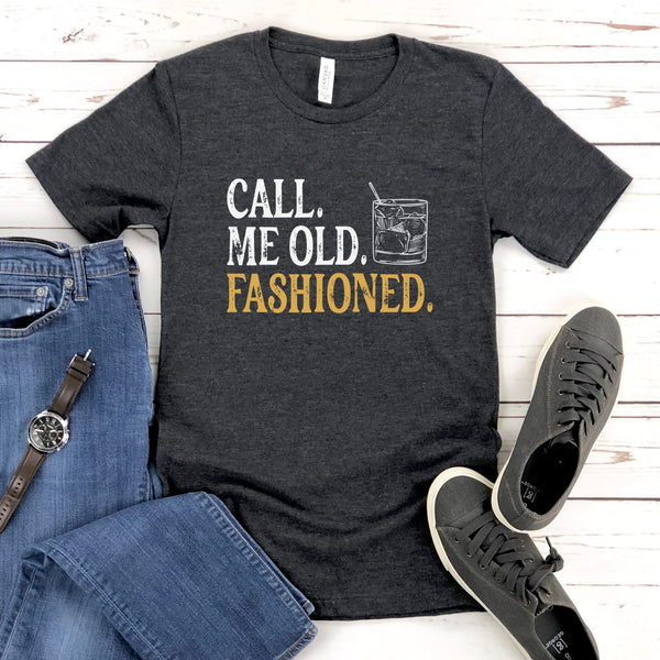 a gray t-shirt with the text "Call me old, fashioned" printed on it, along with a graphic of a cocktail glass. The t-shirt is laid out on a wooden surface, accompanied by a pair of blue jeans, a watch, and a pair of sneakers.