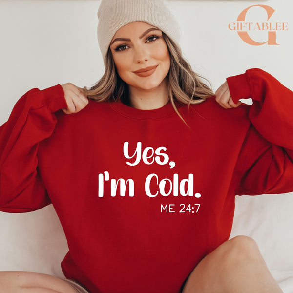 A woman wearing a red sweater with the text "Yes, I'm cold" and "ME 24:7" printed on it.