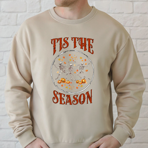 A beige sweater with a Halloween-themed graphic design. The design features a circular arrangement of skeleton figures and pumpkins, along with the text "TIS THE SEASON" written above the design.