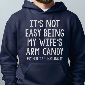 A navy blue hoodie with white text that reads "IT'S NOT EASY BEING MY WIFE'S ARM CANDY BUT HERE I AM NAILING IT".