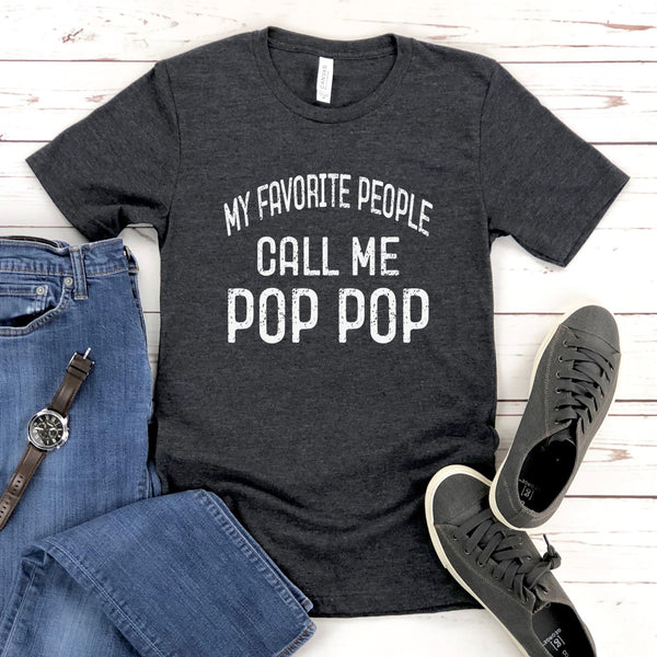 A gray t-shirt with the text "MY FAVORITE PEOPLE CALL ME POP POP" printed on it, along with a pair of jeans and sneakers, all laid out on a wooden surface.
