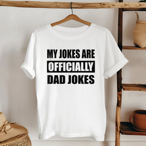 A white t-shirt with the text "MY JOKES ARE OFFICIALLY DAD JOKES" printed on it, hanging on a wooden hanger.