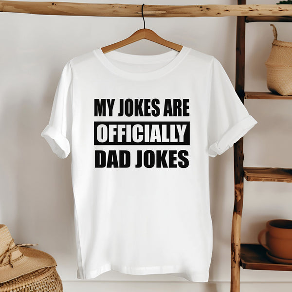 A white t-shirt with the text "MY JOKES ARE OFFICIALLY DAD JOKES" printed on it, hanging on a wooden hanger.
