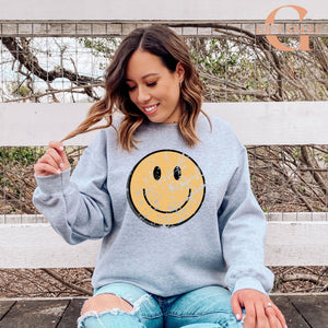 A woman sitting on a bench with a distressed yellow smiley face design on her gray sweatshirt.