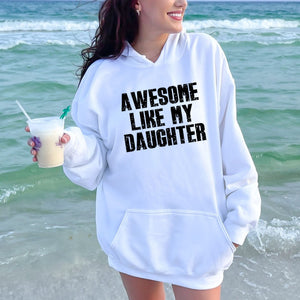 A woman wearing a white hoodie with the text "AWESOME LIKE MY DAUGHTER" printed on it, standing on a beach with the ocean in the background.