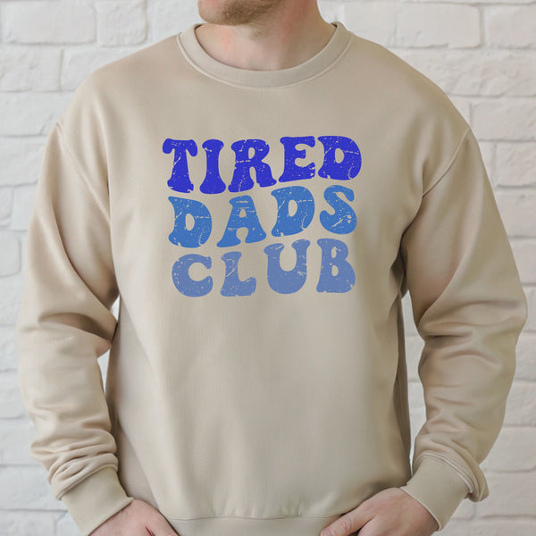 A man wearing a beige sweater with the text "TIRED DAD'S CLUB" printed on it.