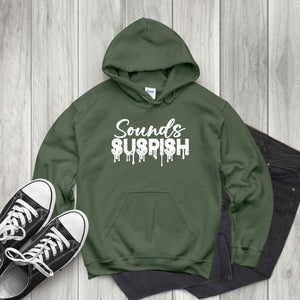 A green hoodie with the text "Sounds Suspect" printed on it, along with a pair of black and white sneakers, is laid out on a wooden surface.