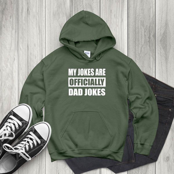 A green hoodie with white text that reads "MY JOKES ARE OFFICIALLY DAD JOKES" is displayed on a wooden surface alongside a pair of black and white sneakers.