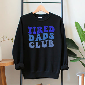 A black sweatshirt with the text "TIRED DAD'S CLUB" printed on it, hanging on a wooden hanger.