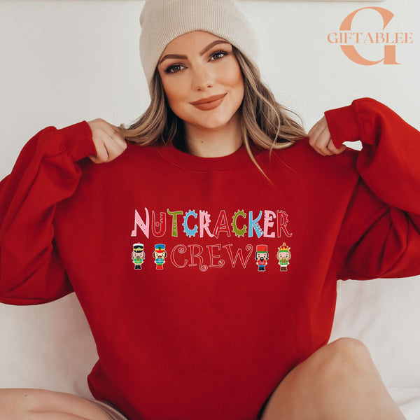 A woman wearing a red sweater with the text "NUTCRACKER CREW" printed on it, smiling and posing with her arms behind her back.