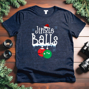 A navy blue t-shirt with a humorous text design that reads "Jingle Balls" and features a Santa hat and Christmas ornaments. The t-shirt is placed on a wooden surface, surrounded by holiday decorations such as pine branches.