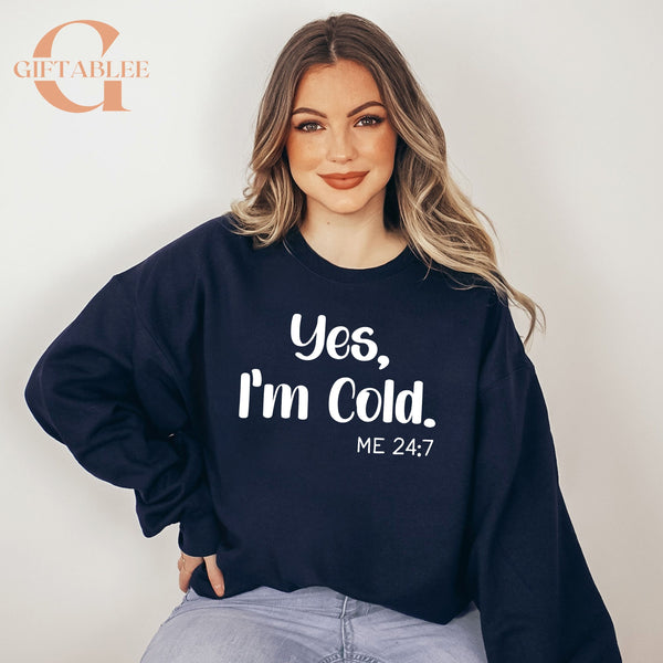 A woman wearing a navy blue sweatshirt with the text "Yes, I'm Cold. ME 24:7" printed on it.