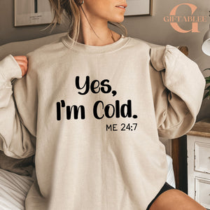 A woman wearing a beige sweatshirt with the text "Yes, I'm Cold. ME 24:7" printed on it.