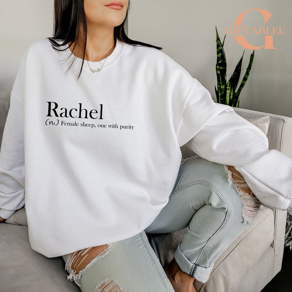 A woman wearing a white sweatshirt with the word "Rachel" printed on it, sitting on a couch.