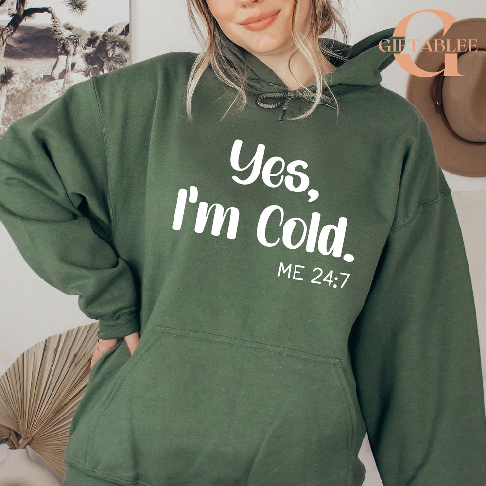 A woman wearing a green hoodie with the text "Yes, I'm Cold. ME 24:7" printed on it.