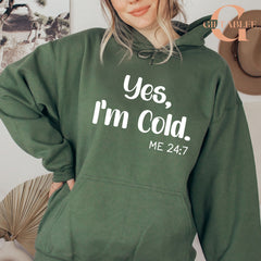 A woman wearing a green hoodie with the text "Yes, I'm Cold. ME 24:7" printed on it.