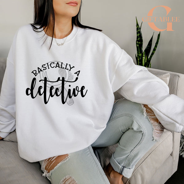 A woman wearing a white sweatshirt with the text "BASICALLY A DETECTIVE" printed on it, sitting on a couch.