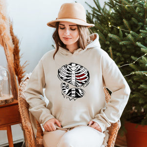 A woman wearing a beige hat and a white hoodie with a graphic of a skeleton on it, sitting in a wicker chair in front of a Christmas tree.