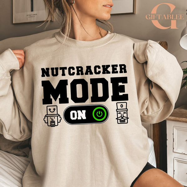 A person wearing a beige sweater with the text "NUTCRACKER MODE ON" printed on it.