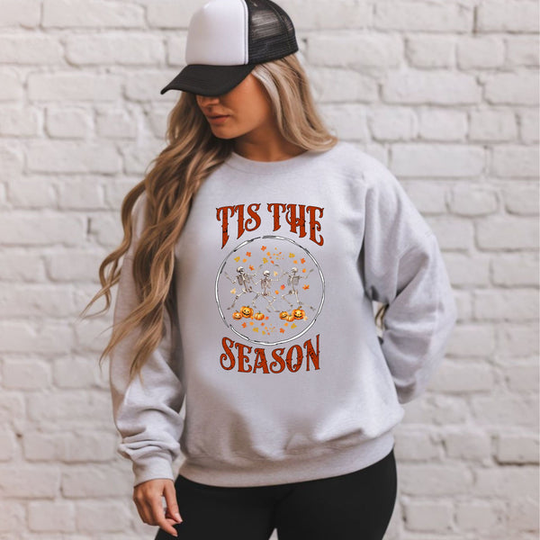 A woman wearing a gray sweatshirt with a graphic design and text printed on it, standing in front of a white brick wall.
