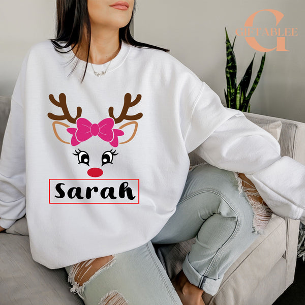 A woman wearing a white sweater with a reindeer face design and the name "Sarah" printed on it, sitting on a couch.