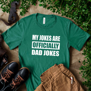 A green t-shirt with the text "MY JOKES ARE OFFICIALLY DAD JOKES" printed on it, along with a pair of brown pants and black shoes, all placed on a surface with some greenery.