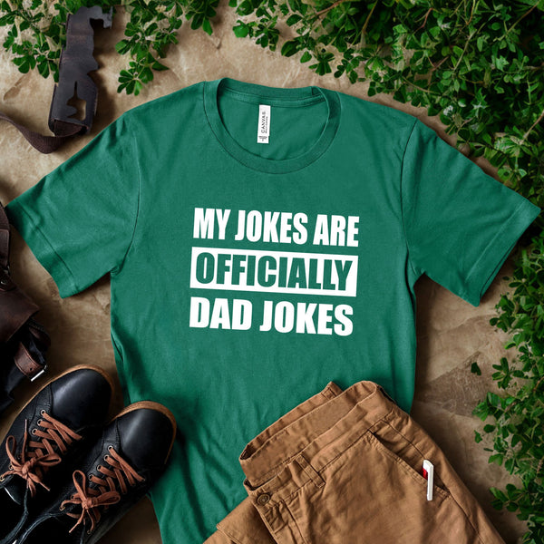 A green t-shirt with the text "MY JOKES ARE OFFICIALLY DAD JOKES" printed on it, along with a pair of brown pants and black shoes, all placed on a surface with some greenery.