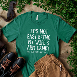 A green t-shirt with a humorous text printed on it, along with a pair of brown pants and black shoes, all placed on a surface with some greenery.