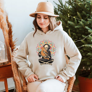 A woman wearing a beige hat and a white hoodie with a graphic of a skeleton holding a guitar is sitting in a wicker chair in front of a Christmas tree.