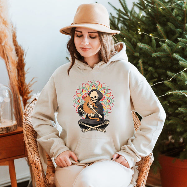 A woman wearing a beige hat and a white hoodie with a graphic of a skeleton holding a guitar is sitting in a wicker chair in front of a Christmas tree.