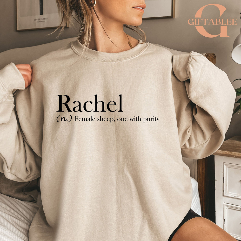 A woman wearing a beige sweater with the word "Rachel" printed on it, sitting on a bed.