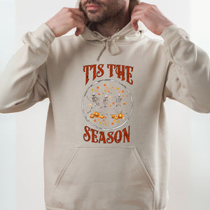 A man wearing a beige hoodie with a graphic design featuring a skeleton and pumpkins, along with the text "TIS THE SEASON" printed on it.