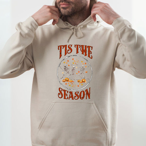 A man wearing a beige hoodie with a graphic design featuring a skeleton and pumpkins, along with the text "TIS THE SEASON" printed on it.