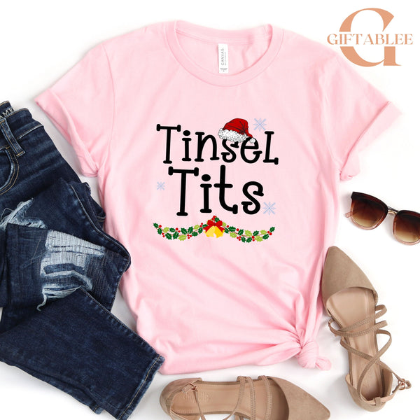 A pink t-shirt with the text "Tinsel Tits" printed on it, along with a Santa hat and Christmas decorations, is displayed alongside a pair of jeans, sunglasses, and a pair of strappy sandals.