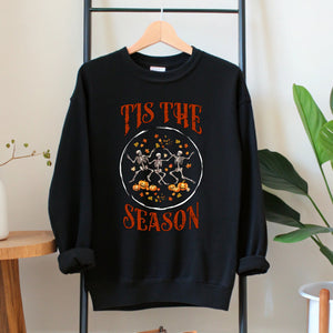 A black sweatshirt with a graphic design featuring three skeletal figures dancing and holding pumpkins, along with the text "Tis the SEASON" printed on it.