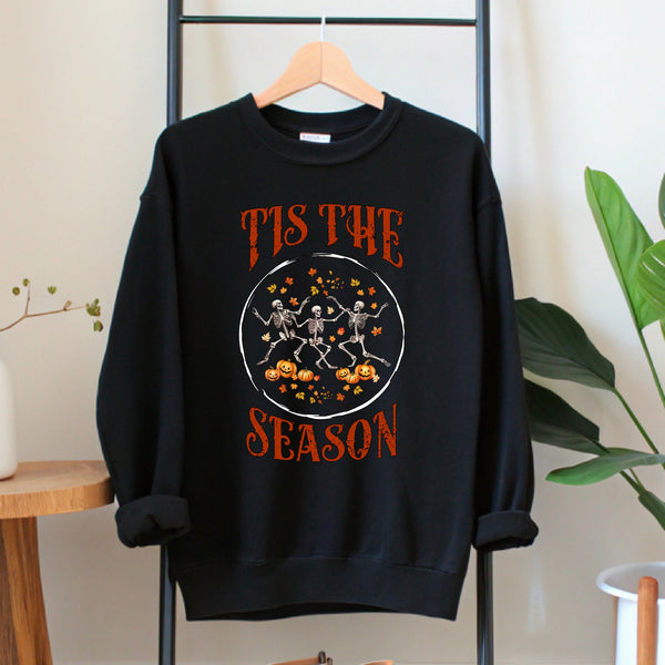 A black sweatshirt with a graphic design featuring three skeletal figures dancing and holding pumpkins, along with the text "Tis the SEASON" printed on it.