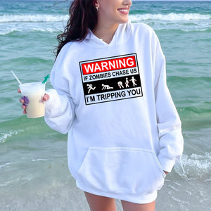 A woman wearing a white hoodie with a humorous warning graphic printed on it, standing near the ocean.