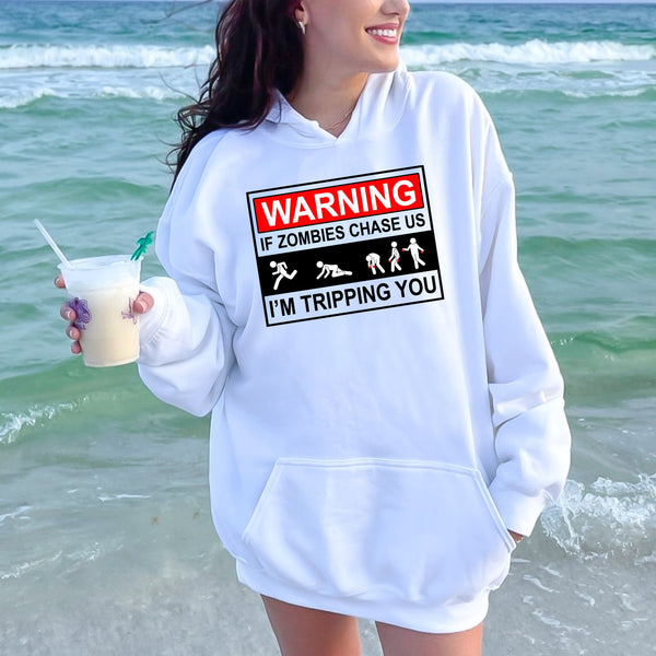 A woman wearing a white hoodie with a humorous warning graphic printed on it, standing near the ocean.
