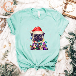 A turquoise t-shirt with a graphic of a pug wearing a Santa hat and Christmas lights, surrounded by holiday decorations such as a framed picture, beads, and greenery.