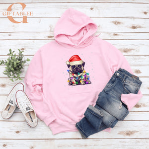 A pink hoodie with a pug wearing a Santa hat, a pair of jeans, and a pair of white sneakers, all laid out on a white wooden surface.
