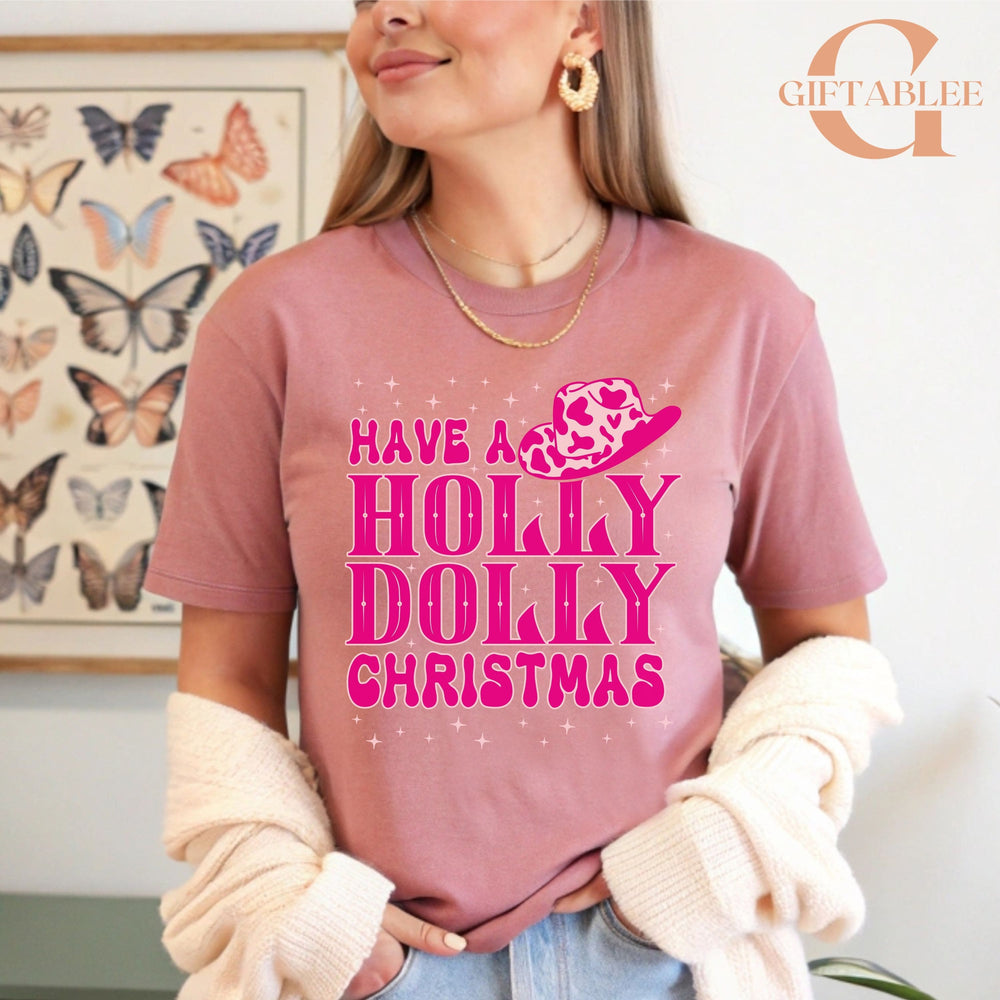 A woman wearing a pink t-shirt with a Christmas-themed design and text, standing in front of a butterfly-themed poster.