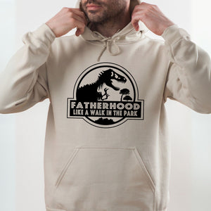 A man wearing a beige hoodie with a graphic design featuring a dinosaur and the text "FATHERHOOD LIKE A WALK IN THE PARK".