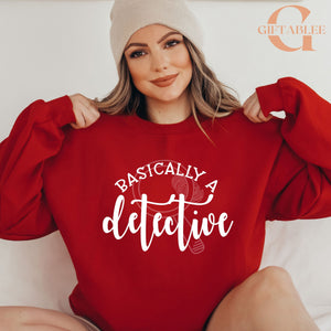 A woman wearing a red sweater with the text "BASICALLY A DETECTIVE" printed on it, smiling and posing with her arms behind her back.