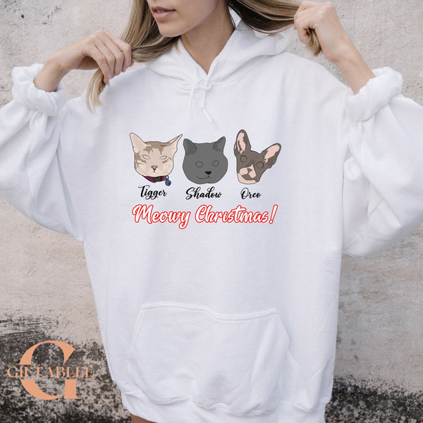 A person wearing a white hoodie with a graphic design featuring three cat faces and the text "Meowy Christmas!"