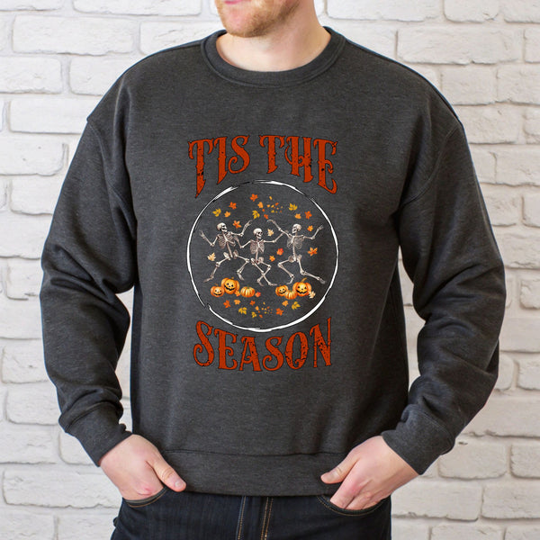 A man wearing a grey sweatshirt with a graphic design featuring three skeletons dancing and a text that says "TIS THE SEASON".