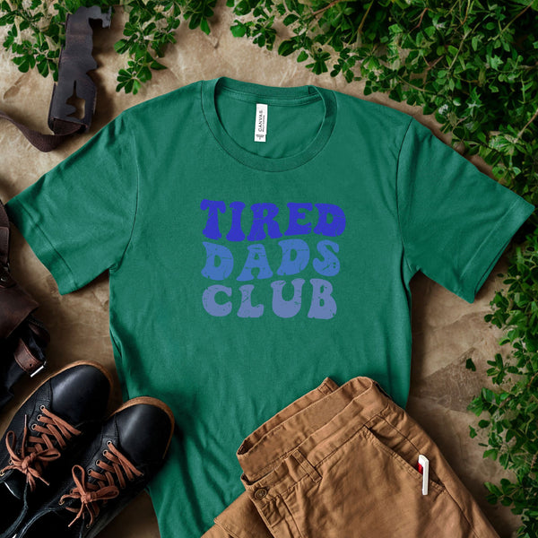a green t-shirt with the text "TIRED DAD'S CLUB" printed on it, along with a pair of brown pants and black shoes. The t-shirt is laid out on a surface with some greenery, such as leaves and branches, creating a casual and relaxed atmosphere.
